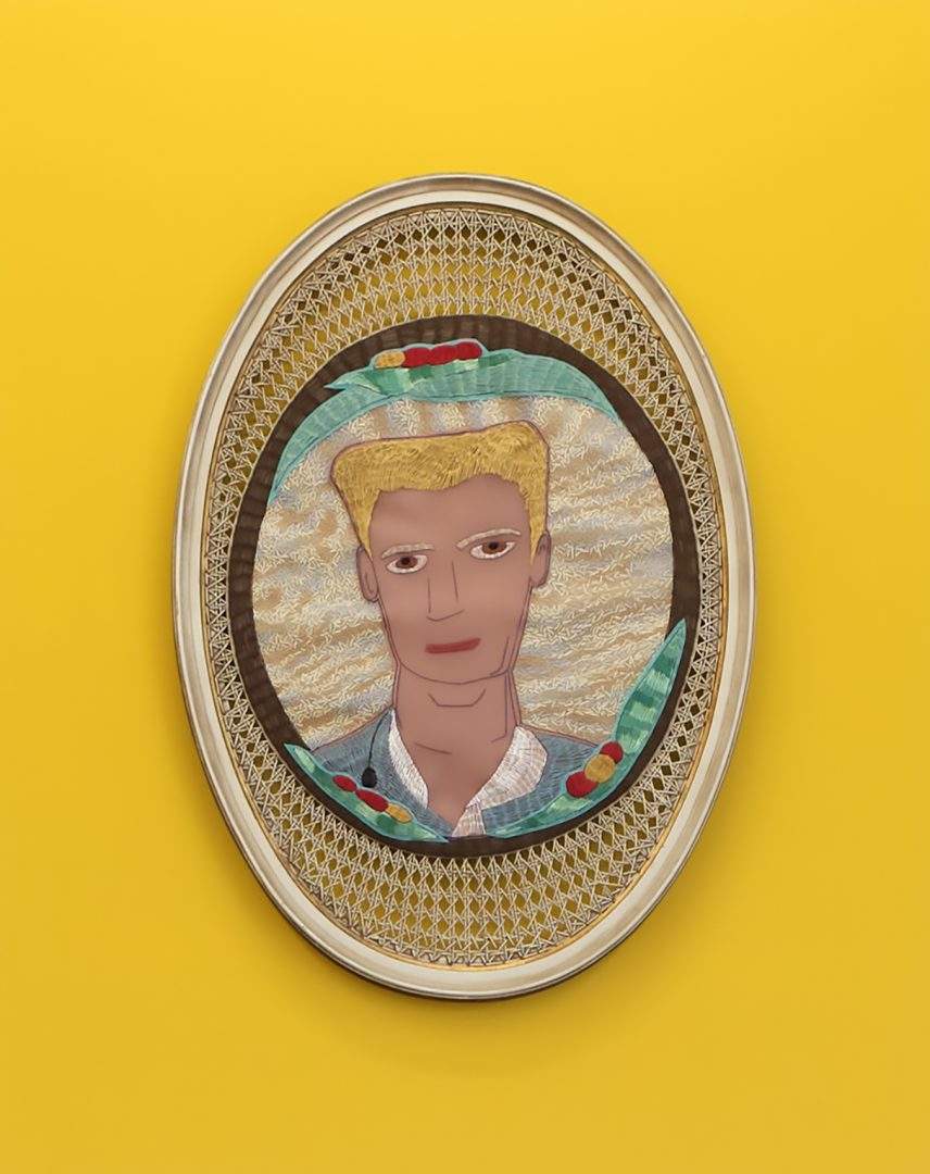 Obra de f.marquespenteado em formato elíptico feita de um cesto com um bordado colado ao centro que representa um homem loiro. A obra está apoiada sobre uma parede amarela.