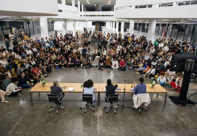Dentro do Pavilhão da Bienal, o coletivo curatorial composto por Manuel Borja-Villel, Diane Lima, Grada Kilomba e Hélio Menezes estão sentados de costas em uma mesa enquanto um público de em torno de 400 pessoas observa atentamente ao coletivo