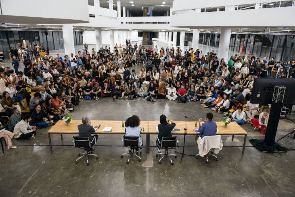 Dentro do Pavilhão da Bienal, o coletivo curatorial composto por Manuel Borja-Villel, Diane Lima, Grada Kilomba e Hélio Menezes estão sentados de costas em uma mesa enquanto um público de em torno de 400 pessoas observa atentamente ao coletivo