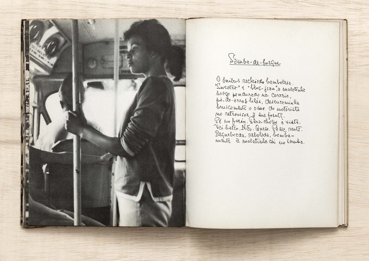 Livro aberto com uma foto em preto e branco de uma pessoa em pé num ônibus segurando-se numa barra, e na página esquerda, um texto escrito a mão onde se lê "Samba-do-brique" e a continuação de um texto.