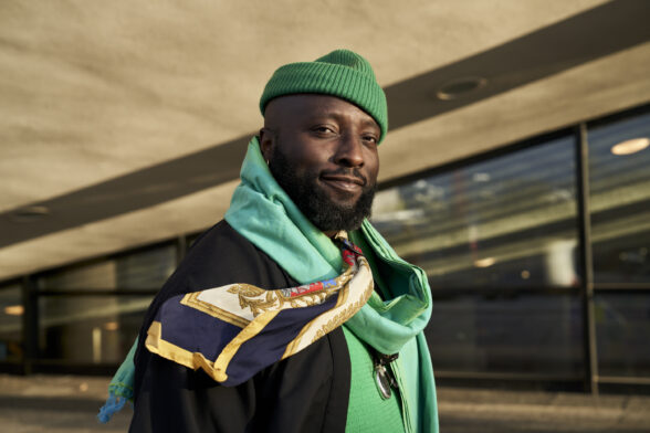 Foto de Bonaventure S. B. Ndikung, que mostra em primeiro plano um homem negro, de barba, usando um gorro verde, lenço estampado, roupa verde com casaco preto, olhando diretamente para a câmera com um leve sorriso, diante de um prédio de concreto e vidro